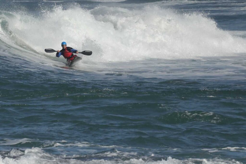 A kayaker rides a large breaking wave, demonstrating challenging water conditions