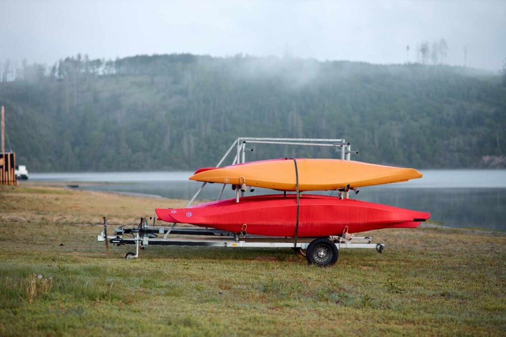 Red and orange kayaks secured on a multi-tier trailer rack near a foggy lake, showing proper loading setup