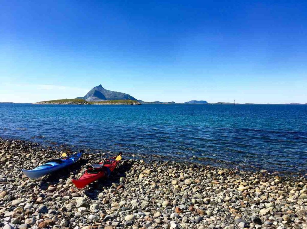 Blue and red kayaks resting on a pebble beach beside clear blue coastal water with a dramatic island peak in the background