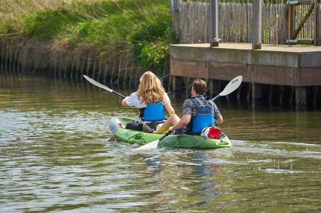 Two people paddling a green recreational tandem kayak on a calm river near a wooden dock