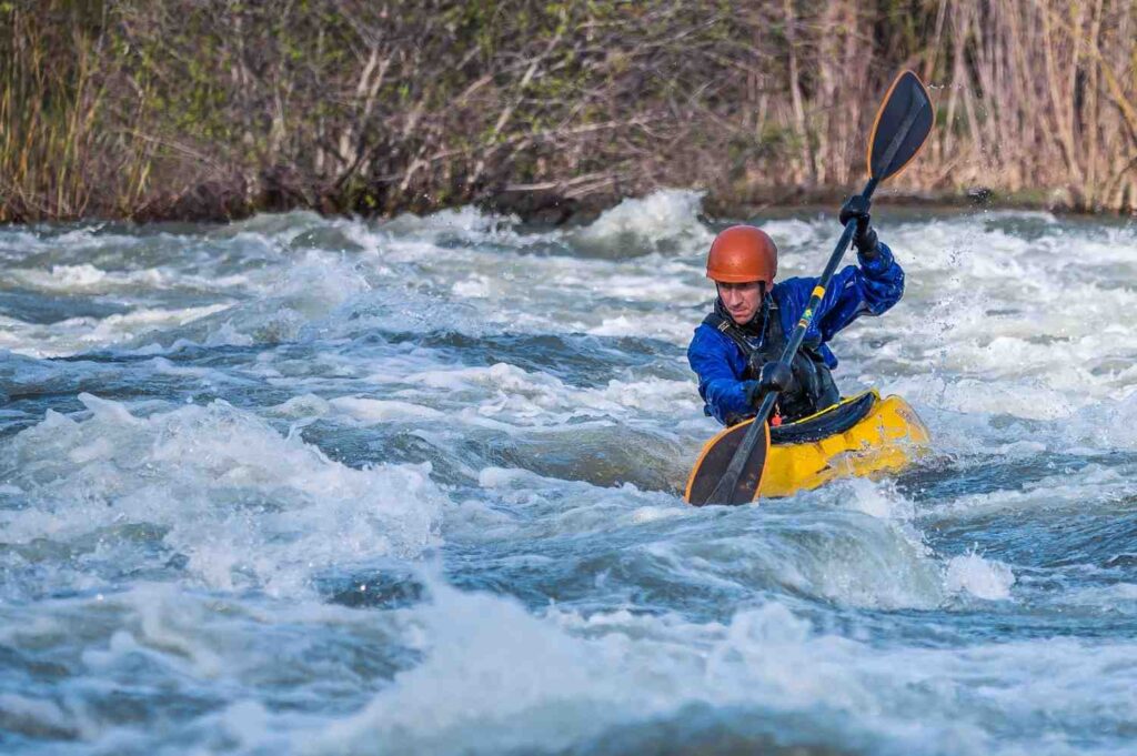 A kayaker wearing a helmet paddles through churning white rapids on a fast-moving river