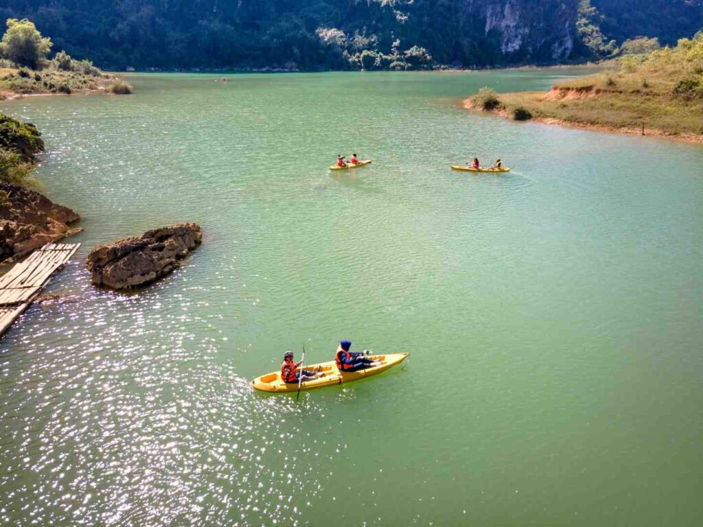 Multiple people paddling wide, stable recreational kayaks on a calm green lake surrounded by cliffs