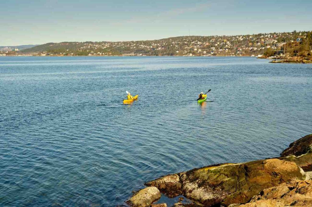 Two people kayaking side by side in yellow and green kayaks near a rocky shoreline with a town in the distance