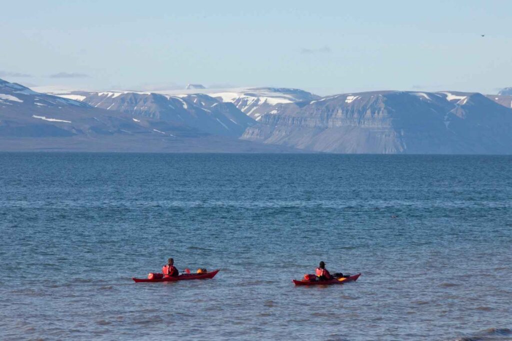 Two red touring kayaks with camping gear paddling across a calm open bay with mountains in the background